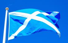 The flag of Scotland - a white cross on a blue background - flutters on the top of a flag post in bright sunshine