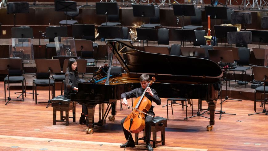 Two children on stage, one playing cello and the other playing a Steinway grand piano