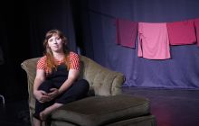 Performer sitting on a chaise longue, looking out, deep in thought. Background is dark, lit to focus on the performer and her laundry hanging behind (visual red flags).