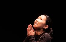 Actor Zoe Kim sits on the floor, palms in prayer, looking up. 
