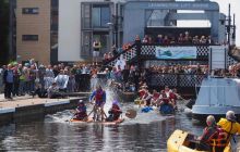 Boaters on strange vessels compete in a raft race on the Union Canal with spectators crowded on the Leamington Lift Bridge behind them