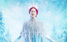 Claire Dargo as the Snow Queen, faces the camera wearing a blue ballgown with a beaded shawl that fastens in the front. She wears long, light blue gloved and a large sparkly crown sits upon her red hair. Diamonds cover her eye lids, and the backdrop is of a wintery blue forest. The Storytelling PR logo sits in the bottom centre of the frame.