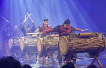 Drummers in a line, lit by blue light, with white beams highlighting