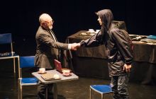 Two men greet each other with a handshake. One is dressed in a suit, the other in a rain coat with a hood.