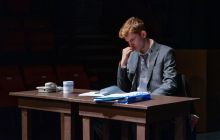 The lead actor as The Lawyer, sits at a table looking at papers, with a thoughtful look on his face.