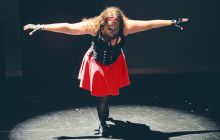 Performer Torrey Shine, dressed in black bustier, pink skirt, looks down with her arms outstretched, lit in a circle, but surrounded by darkness