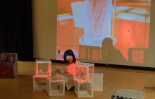 Performer Xiner Lan sits on the floor in front of white and orange boxes, with a projection screen in the background, shown with an orange wash