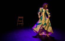 Performer Maria MacDonell starts, highlighted in a dark background, in a long glamorous cream coloured robe that has large bright blue peacocks on it.