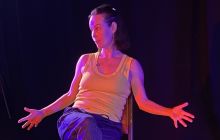 Performer Ambrosine Davies sits in a chair, with her arms extended, in dark light with a spotlight. She wears dark blue jogging trousers and a lime green vest.