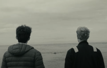 Two young men stand and look out at the wide open sea. The day seems grey and gloomy.