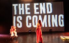 Screen with the words 'the end is coming' angled across it. Performance artist Louise Orwin stands in front of it with her back to the audience, wearing a red strappy top and red tracksuit bottoms. A giant teddy bear is seated to the left of the screen.