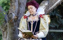 Elizabeth in her wig and full costume, looking at a book with her head tilted.