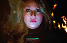 Woman looks directly at the screen. Darkness is behind with some fire. Fierce look.
