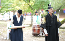A courtier stands to the left of the King, with the Queen in the background, pictured in a green leafy area.