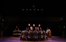 Drummers are pictured with their drums, in front of a dark background.