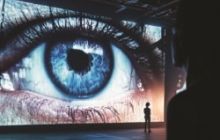 An eye watches over you. A person stands in front of it, very small in comparison.