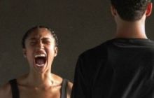 A woman screams with her eyes shut. A man stands with his back to the camera.