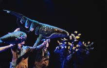 Whale puppet moves, held by puppeteers. Dark background, whale is lit up with lights showing on head.
