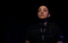 PC Irie looks out into the world, a serious look on her face. There is darkness around her but she is in the spotlight.