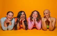 The four members of the girl group lie on the ground, with their faces on their crossed arms. Background is a mustard yellow. All are smiling.