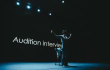 Hyojin Park stands in front of a screen on which the words 'Audition Interview' appear. The background is dark with the performer standing with arms outstretched in silhouette.