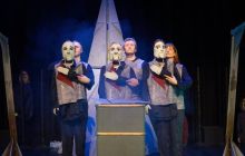 Performers on stage with the puppet masks, stand in the centre, large item at the back behind them