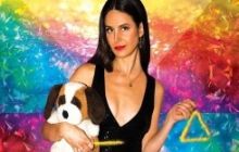 Brooke in a black top and trousers, holding a toy dog, with a rainbow coloured background.