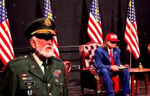 The General stands on the left, Trump is in the middle and several American flags surround the space.