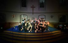 Large cast photo pose with arms out, black background, all seen in the spotlight. Church background shown.