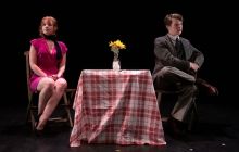 The two actors are pictured on stage, the female Comrade to the left, the male Agent on the right, with a dinner table between them, covered in a red and white checked table cloth.