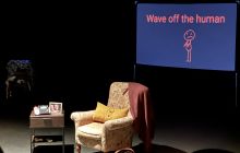 A screen shows in the background, an armchair and small bedside table to the side. The lighting is dark, with the furniture lit.