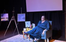 Performer/playwright Patricia McGrath sits next to a screen. There is dark blue light and paintings to the side. Everything is awash with shades of blue.