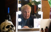 Nick Ullett, the actor and writer, is reflected in a dressing room mirror, lights around it, and a skull to his left.