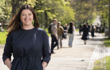 Book Festival director, Jenni Niven, wearing a black summer dress, smiles to the camera on Middle Meadow Walk. In the background, are walkers passing up the hill.