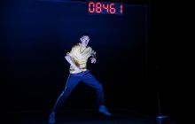 A male dancer is lit in the centre of a dark space with a screen showing numbers in red in the background