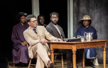 Court scene with Richard Coyle as Atticus Finch, Aaron Shosanya as Tom Robinson seated at a table.