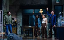 Four people stand in a room, three with their hands in the air, looking at the fourth,