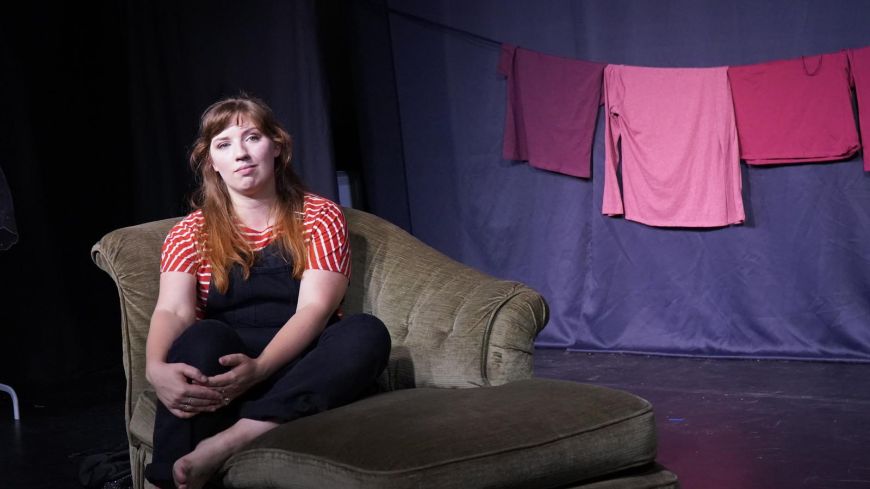 Performer sitting on a chaise longue, looking out, deep in thought. Background is dark, lit to focus on the performer and her laundry hanging behind (visual red flags).