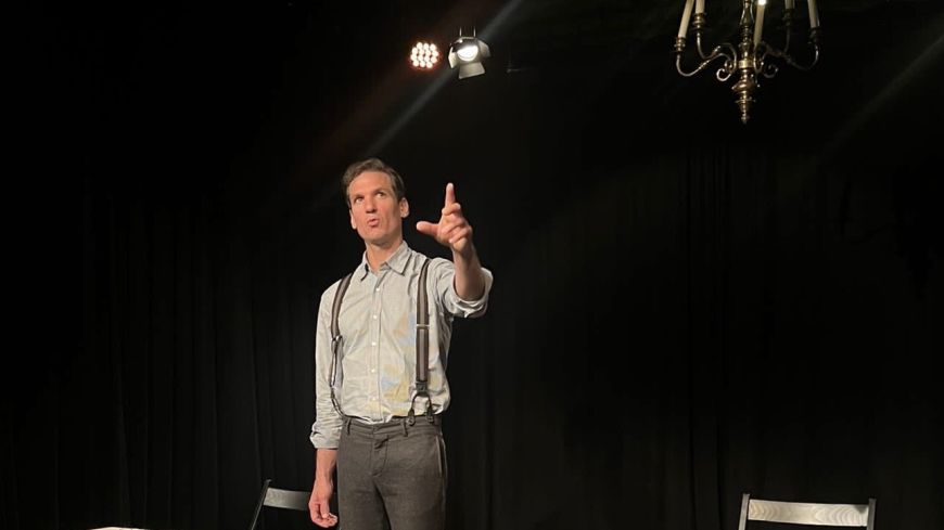 Actor Luke Townson stands looking out, with a background of a black box theatre space, chairs and books visible