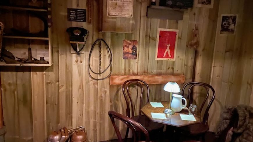 A darkened room with a table, chairs, and wooden walls. Teapot and glasses on the table.