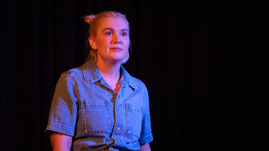 Writer/Performer Kirby Lunn is pictured in a dark background, looking up, wearing a denim jumpsuit