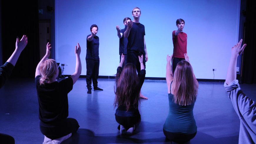 A white and blue lit backdrop highlights the outline of performers in lines, arms reaching up, from the show Death by Shakesepeare