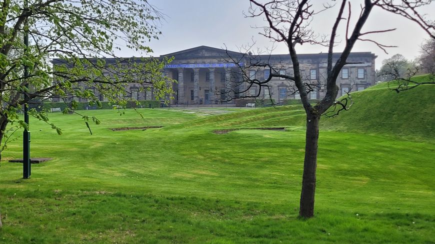 Modern art gallery appears through saplings and grassy, undulating park of landform