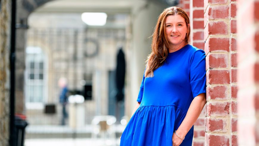 Edinburgh International Book Festival Jenny Niven in a blue dress leans casually against a brick wall 