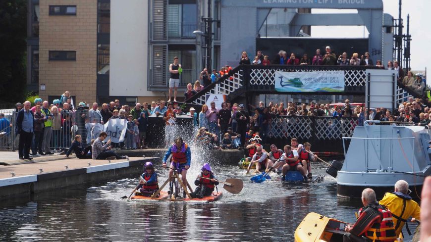 Boaters on strange vessels compete in a raft race on the Union Canal with spectators crowded on the Leamington Lift Bridge behind them