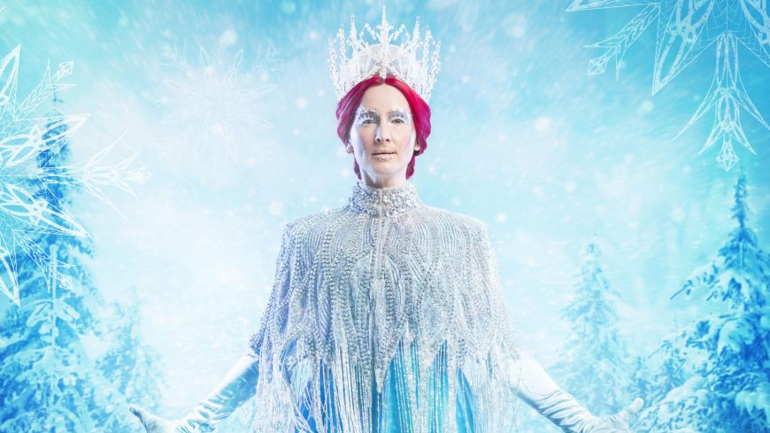 Claire Dargo as the Snow Queen, faces the camera wearing a blue ballgown with a beaded shawl that fastens in the front. She wears long, light blue gloved and a large sparkly crown sits upon her red hair. Diamonds cover her eye lids, and the backdrop is of a wintery blue forest. The Storytelling PR logo sits in the bottom centre of the frame.