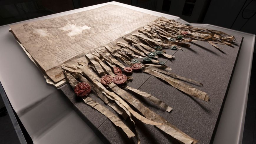 A worn and aged document with medieval seals strung flimsily from it is spread out in a display cabinet in low light. This is the Declaration of Arbroath