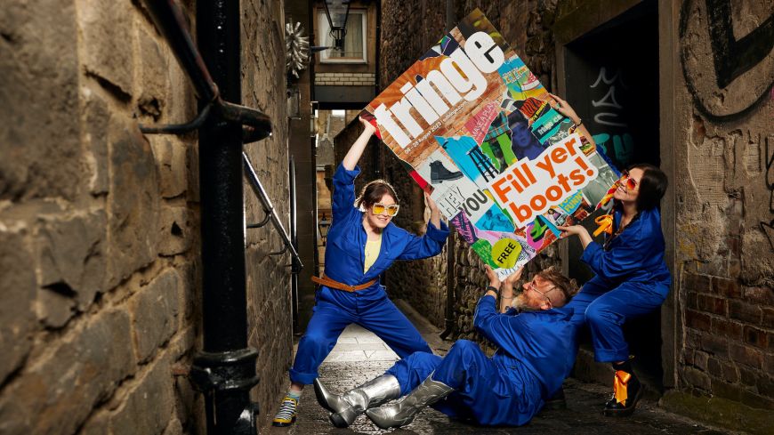 Three models in blue overalls (Claudia Cawthorne, Martha Haskins, Chris Peploe) hold up a large copy of the Fringe 2023 brochure in a narrow alleyway in Edinburgh's Old Town. On it are the words "Fill Yer Boots."