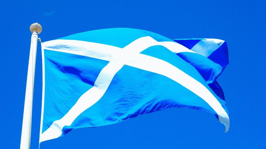 The flag of Scotland - a white cross on a blue background - flutters on the top of a flag post in bright sunshine