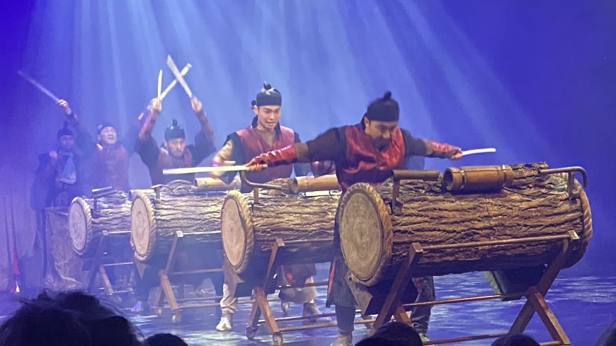 Drummers in a line, lit by blue light, with white beams highlighting